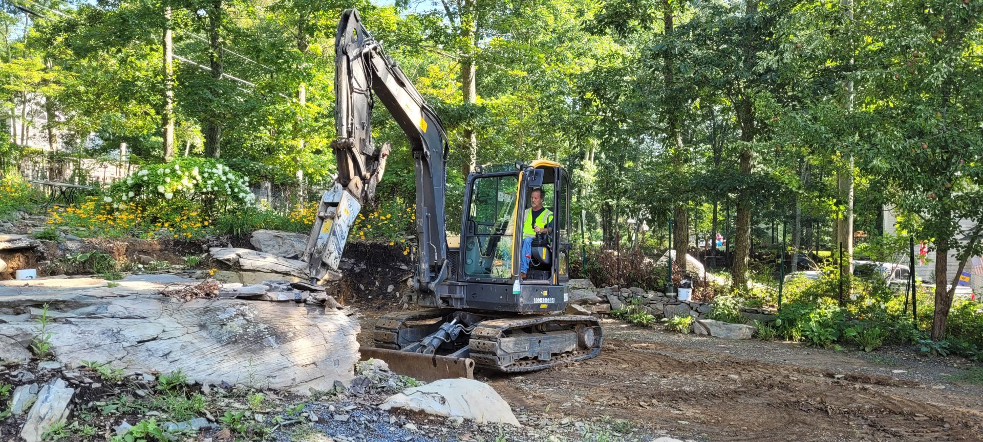 A yellow backhoe working on a personal property to level the ground for landscaping, with piles of dirt and rocks