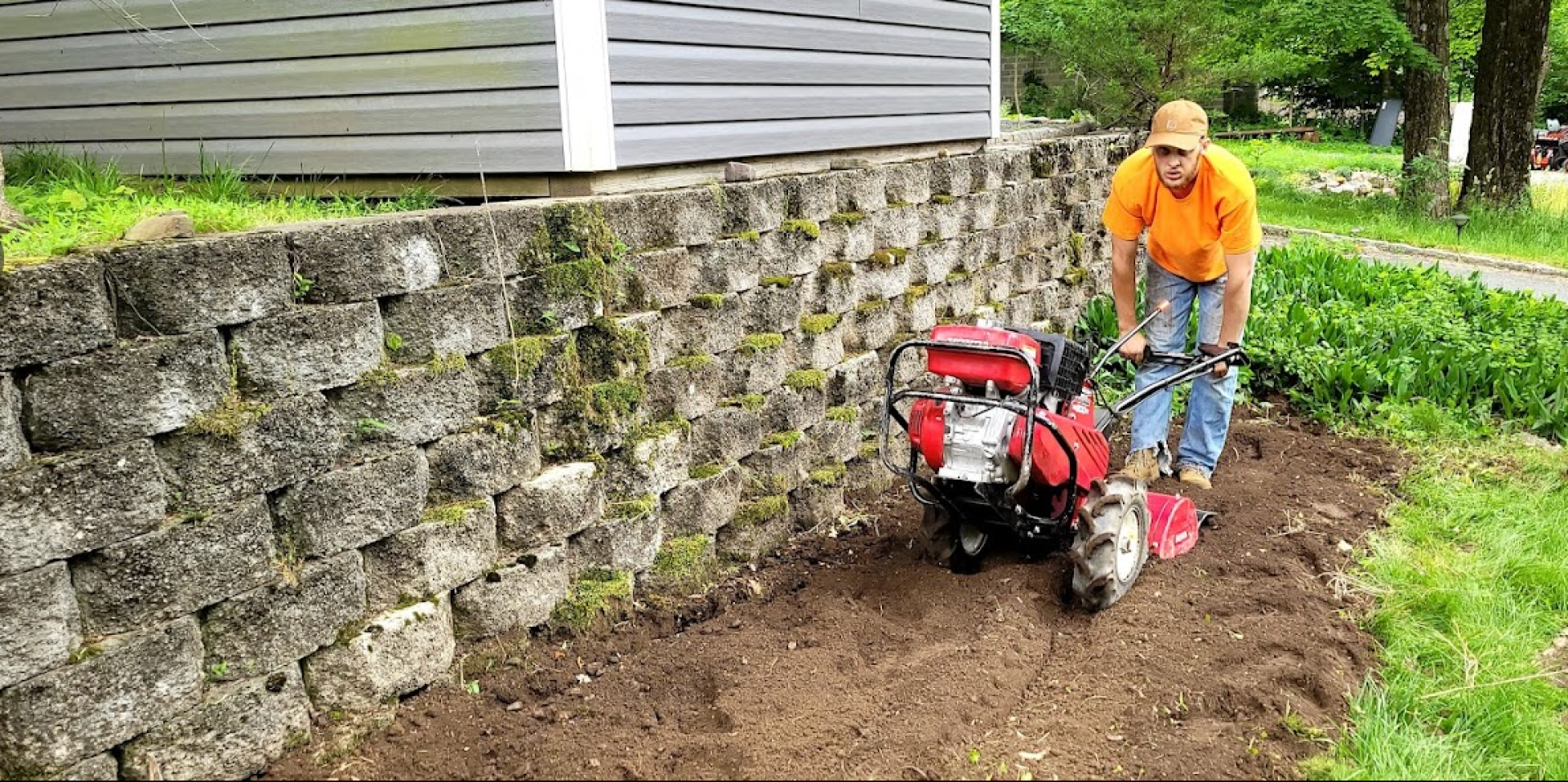 A man in an orange shirt tilling dirt with a red machine alongside a retaining wall made of stacked gray bricks