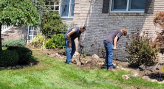 Two men in a yard working on landscaping, both shoveling in garden bed area near brick home